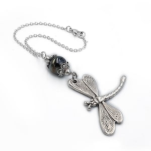 Könnte beinhalten: Ein silberfarbener Libellenanhänger mit detaillierten Flügeln und einem gebogenen Schwanz hängt an einer zarten Kette. Über der Libelle befindet sich eine dunkle, gemusterte Perle. Die Kette hat oben einen Verschluss.