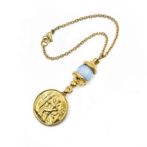 Könnte beinhalten: Ein goldfarbenes Kettenarmband mit einem runden, geprägten Münzanhänger, der eine Figur darstellt, sowie einer einzelnen, glatten, hellblauen Kugelperle, die von verzierten goldfarbenen Kappen gehalten wird. Mit Karabinerverschluss.