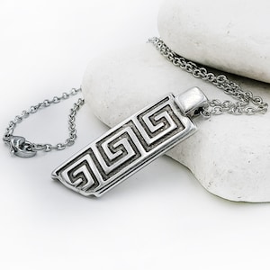 Op de afbeelding: Een zilveren ketting met een rechthoekige hanger met een Grieks sleutelmotief. De hanger hangt aan een zilveren ketting.