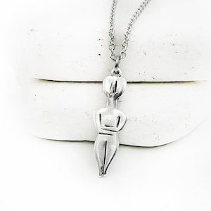 Op de afbeelding: Een zilverkleurige ketting met een hanger in de vorm van een gestileerde vrouwenfiguur. De hanger heeft een minimalistisch ontwerp met een matte afwerking en hangt aan een fijne ketting. De ketting wordt gepresenteerd tegen een witte achtergrond.
