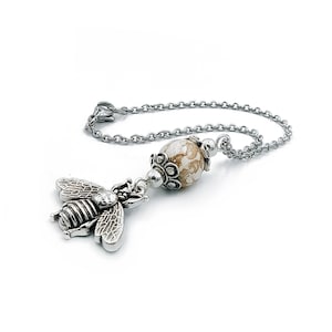 Puede incluir: Un collar de cadena de plata con una cuenta de color beige y blanco con estampado y un dije de abeja de plata.