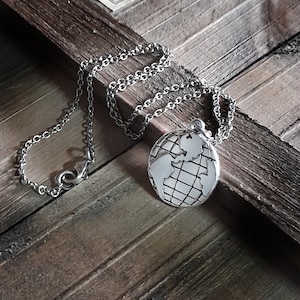 Puede incluir: Un collar de plata con un colgante de globo terráqueo. El globo está detallado con líneas que representan los continentes y un patrón de cuadrícula.