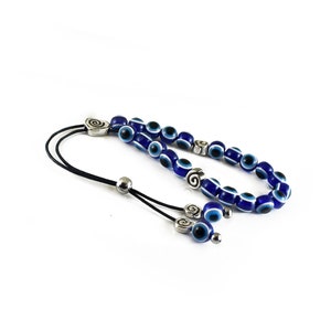 May include: A blue and white evil eye bead necklace with silver accents. The necklace is made of small round beads with a blue background and a white eye design. The necklace has a silver clasp and a silver charm with a spiral design.