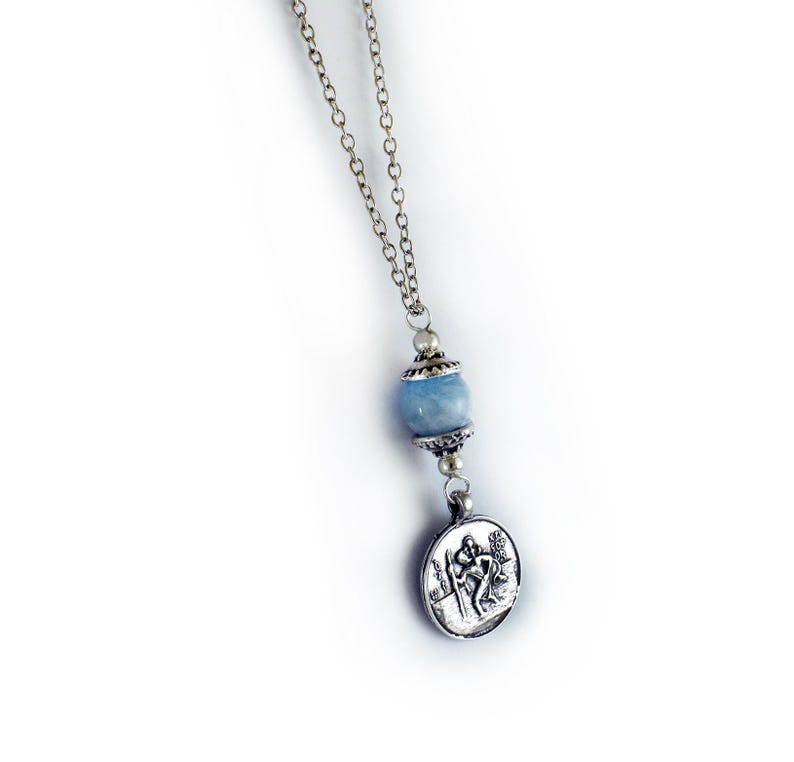 Puede incluir: Un collar de cadena de metal plateado con una cuenta de piedra redonda de color azul claro acentuada por tapas decorativas plateadas. Un colgante circular plateado cuelga de la cuenta, mostrando una imagen en relieve de una figura con un bast&oacute;n.