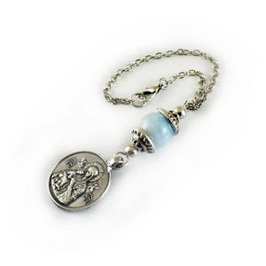 Puede incluir: Una cadena de metal plateado con una cuenta redonda, lisa y de color azul claro, acentuada con tapas decorativas de metal plateado a ambos lados, unida a un colgante ovalado de metal plateado que representa una figura religiosa en relieve.