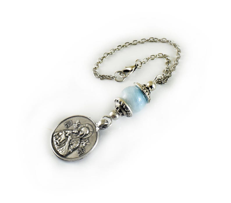 Puede incluir: Una cadena de metal plateado con una cuenta redonda, lisa y de color azul claro, acentuada con tapas decorativas de metal plateado a ambos lados, unida a un colgante ovalado de metal plateado que representa una figura religiosa en relieve.