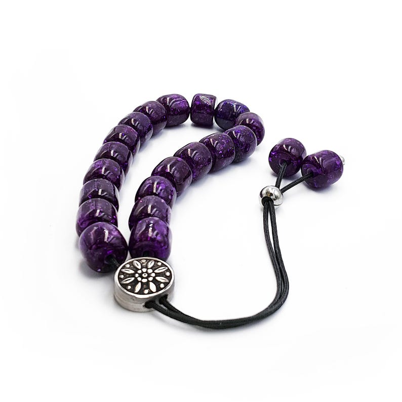 May include: A purple glass prayer bead mala with a silver charm. The beads are round and have a speckled appearance. The mala is strung on a black cord with a silver tassel.