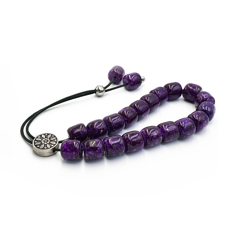 May include: A purple glass prayer bead mala with a silver tassel and a silver charm. The beads are round and have a speckled texture.