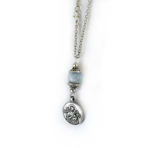 Puede incluir: Un collar de cadena de metal plateado con una cuenta de piedra redonda de color azul p&aacute;lido acentuada con tapas decorativas plateadas. Un colgante circular plateado cuelga de la cuenta, mostrando una imagen en relieve de una figura con un ni&ntilde;o.