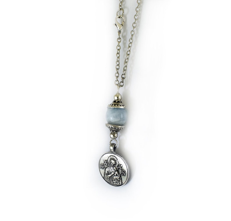 Puede incluir: Un collar de cadena de metal plateado con una cuenta de piedra redonda de color azul p&aacute;lido acentuada con tapas decorativas plateadas. Un colgante circular plateado cuelga de la cuenta, mostrando una imagen en relieve de una figura con un ni&ntilde;o.
