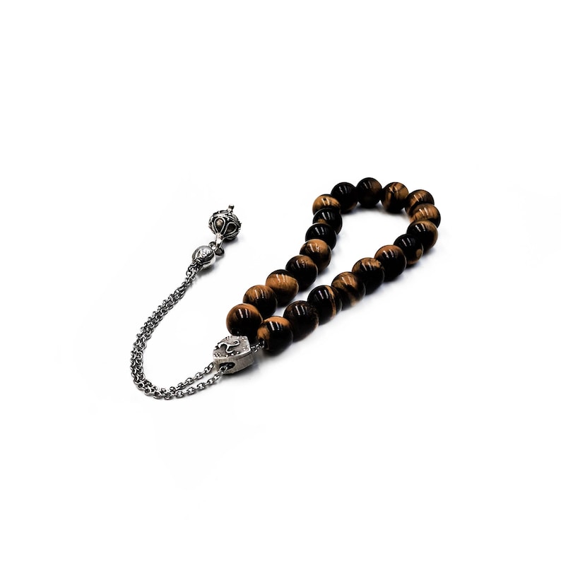 K&ouml;nnte beinhalten: Eine braune und schwarze Tigerauge-Perlenmala mit einem silbernen Quastenanh&auml;nger und einem silbernen Anh&auml;nger. Die Mala ist auf einer silbernen Kette aufgef&auml;delt.
