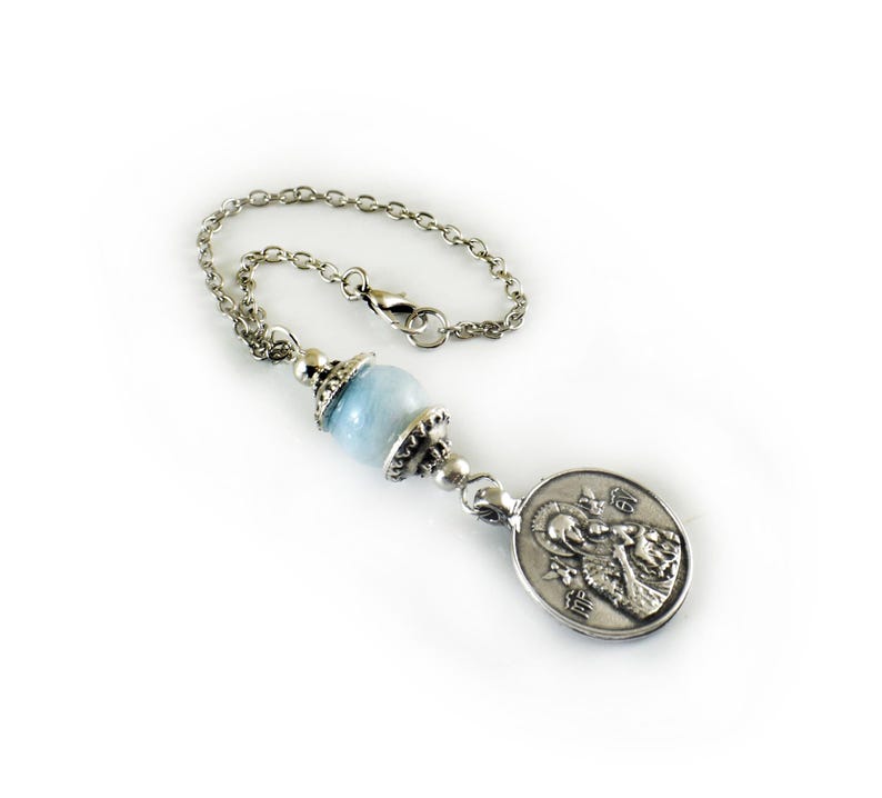 Puede incluir: Una cadena de metal plateado con una cuenta de piedra esf&eacute;rica de color azul claro, acentuada por tapas decorativas plateadas. En el extremo de la cadena hay un colgante de metal plateado de forma ovalada que representa una figura religiosa.