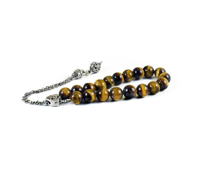 K&ouml;nnte beinhalten: Eine braune und goldene Tigerauge-Perlenmala mit silbernen Akzenten. Die Mala hat einen Quasten und einen silbernen Anh&auml;nger.