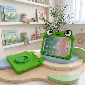 Puede incluir: Una funda de silicona para tablet de color verde brillante con forma de rana, ojos grandes y boca sonriente. Una funda está de pie sosteniendo una tablet con pantalla colorida, mientras que otra funda idéntica y vacía yace plana con un soporte.