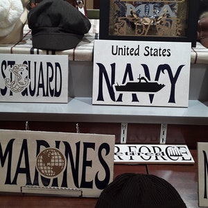 Puede incluir: Una colección de letreros de madera con los nombres de las diferentes ramas de las fuerzas armadas de los Estados Unidos. Los letreros están pintados de blanco con letras negras y presentan varios símbolos, incluyendo un ancla, un barco y un globo terráqueo. Los letreros están dispuestos en una fila en un estante.