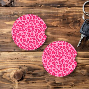 May include: Two pink car coasters with a leopard print design. The coasters are round with a small notch on one side. The background is a wooden surface, and a set of keys is visible.