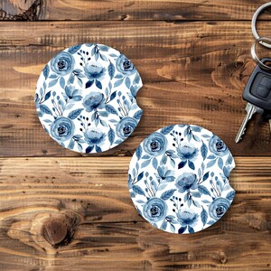 May include: Two car coasters with a floral design. The coasters feature a white background with blue watercolor roses and leaves. A set of car keys is also visible on a wooden surface.