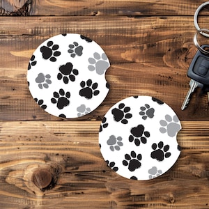 May include: Two white car coasters with a black and gray paw print pattern. The coasters are round with a curved cutout. A set of keys with a black fob and silver ring are in the background.