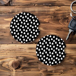 May include: Two black car coasters with a white ghost pattern. The coasters are round with a curved cutout for easy removal. A set of car keys with a black fob and silver ring are also in the image.