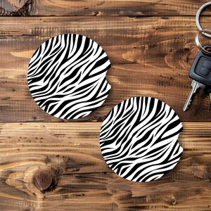 May include: Two round car coasters with a black and white zebra stripe pattern. The coasters have a small notch for easy removal from the cup holder. Keys and a key fob are in the background.