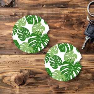 May include: Two round car coasters with a white background and a green monstera leaf pattern. The coasters are on a wooden surface with car keys in the background. The coasters are designed to fit in a car's cup holder.