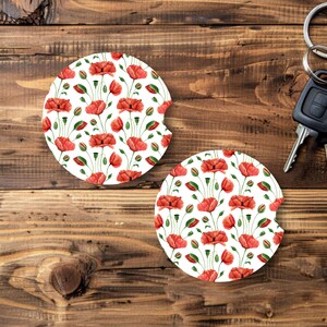 May include: Two white car coasters with a vibrant red poppy floral pattern. The coasters feature a circular design with a curved cutout for easy removal. A set of car keys rests on a wooden surface.