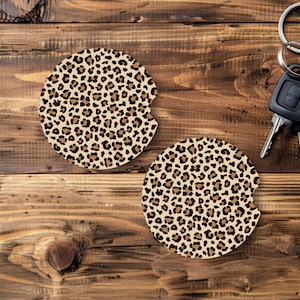 May include: Two round car coasters with a leopard print design. The coasters are beige with brown spots and have a small notch for easy removal. A set of car keys with a black fob and silver ring are also in the image.