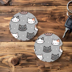 May include: Two gray car coasters with a repeating pattern of cartoon cat faces in white, gray, and orange. The coasters have a small cutout for easy removal. A set of car keys with a black fob and silver ring are in the background.