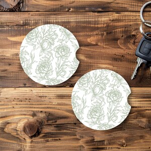 May include: Two white car coasters with a floral design in light green. The coasters have a circular shape with a curved cutout on one side. The background is a wooden surface. Car keys are visible in the background.