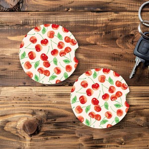 May include: Two round car coasters with a cherry pattern. The coasters feature a cream background with watercolor-style red cherries and green leaves. A car key and keys are in the background.