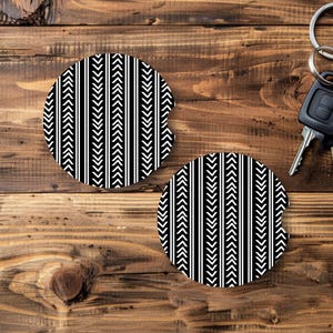 May include: Two black and white car coasters with a geometric arrow and stripe pattern. The coasters are round and made of absorbent material. Car keys are visible in the background.