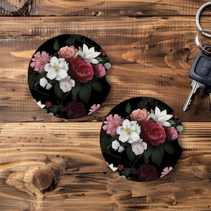 May include: Two black car coasters with a floral design featuring pink, white, and red roses and other flowers. The coasters are on a wooden surface, next to a set of car keys.