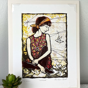 May include: A colorful linocut print of a woman sitting with a red and yellow patterned dress and a red headband. She is surrounded by paper origami birds and the text "It is now" and "Wonders" is visible at the bottom of the print.