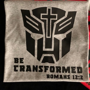 May include: A gray t-shirt with a black Transformer robot graphic with a cross in the middle. The text "BE TRANSFORMED ROMANS 12:2" is printed below the graphic.