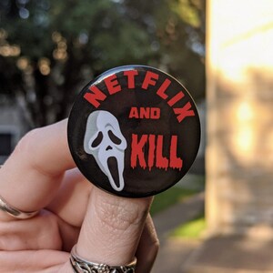 Könnte beinhalten: Ein schwarzer Knopf mit dem Text "Netflix and Kill" in roten Buchstaben. Eine weiße Geistermaske befindet sich in der Mitte des Knopfes.