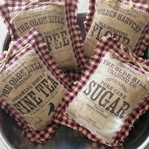 May include: Four fabric sachets with vintage-style labels, nestled in a metal tray. The labels read "Coffee," "Flour," "Tea," and "Sugar." Each sachet is wrapped in a red and cream gingham fabric and tied with twine. A rustic, country-style theme.