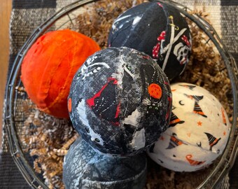 May include: A black wire basket filled with brown paper shreds and four Halloween-themed fabric ornaments. The ornaments are round and feature various patterns, including black cats, witches, and skulls. One ornament is orange.