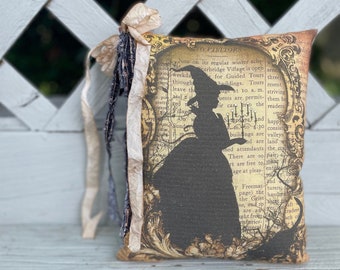 May include: A decorative pillow featuring a silhouette of a witch in a hat and dress, set against a vintage map background. The pillow has a cream-colored ribbon and fabric accents. The text on the pillow reads "TO VISITORS".