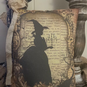 May include: Decorative pillow with a black silhouette of a witch on a vintage book page background. The pillow is square and has a distressed, antique look, perfect for Halloween decor.