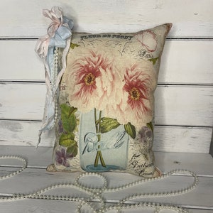 May include: A decorative pillow with a floral print featuring pink peonies in a glass jar. The pillow has a white background with the text "Fleur de Peony Paris" and "Gatherer Peony" printed on it. The pillow is adorned with a blue and pink ribbon and a string of white pearls.