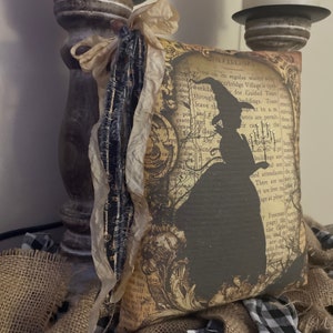 May include: A decorative pillow with a vintage-style design featuring a black silhouette of a witch. The pillow has a cream-colored ribbon and is surrounded by burlap and black and white checkered fabric. The text on the pillow reads "VISITORS".