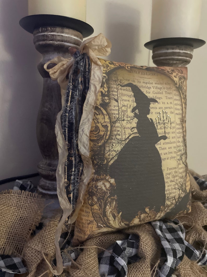 May include: A decorative pillow with a vintage-style design featuring a black silhouette of a witch. The pillow has a cream-colored ribbon and is surrounded by burlap and black and white checkered fabric. The text on the pillow reads "VISITORS".