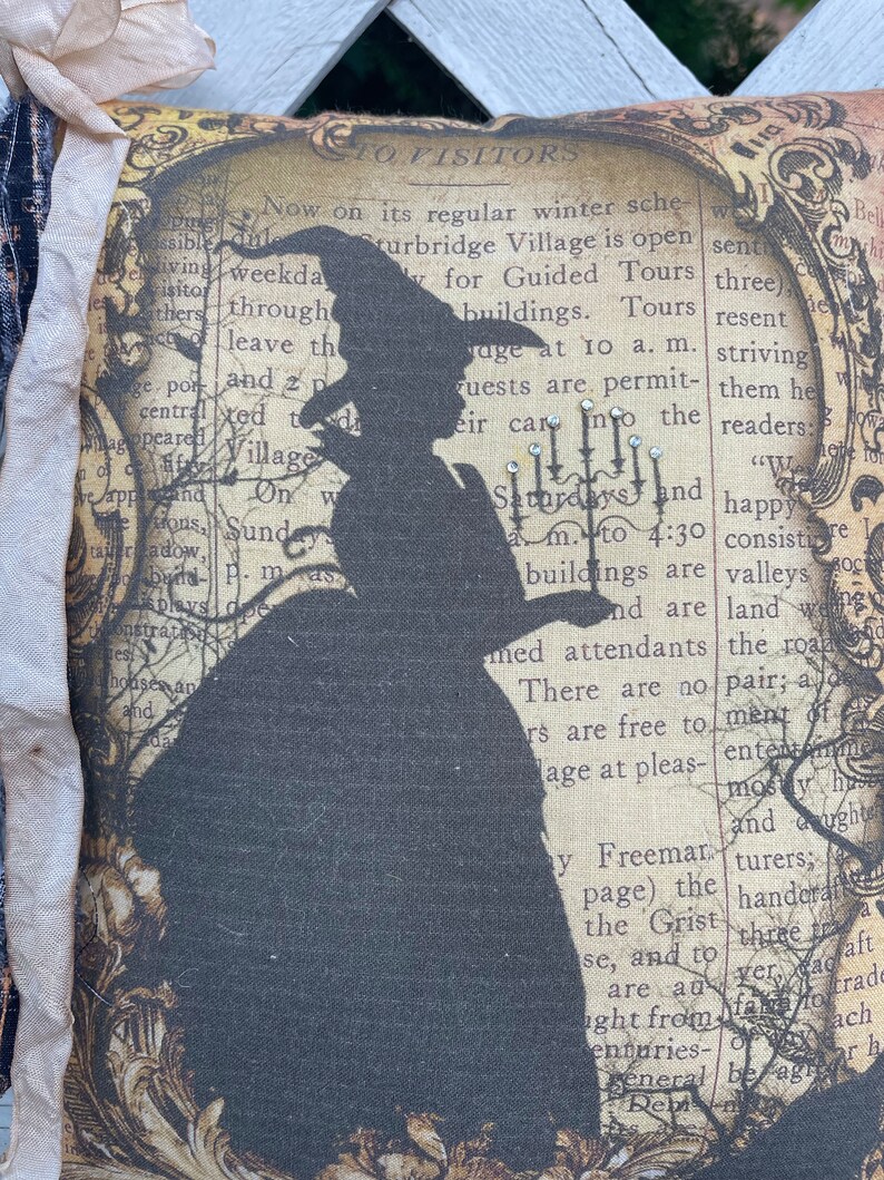 May include: A decorative pillow featuring a silhouette of a witch in a hat and dress, set against a vintage newspaper print background. The pillow has a cream-colored border and is embellished with fabric accents. The design evokes a Halloween or gothic theme.