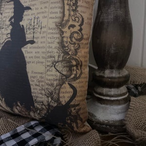 May include: A decorative pillow with a vintage aesthetic, featuring a black silhouette of a witch holding a candle. The pillow has a background of aged paper with text and is surrounded by burlap and a black and white checkered ribbon.