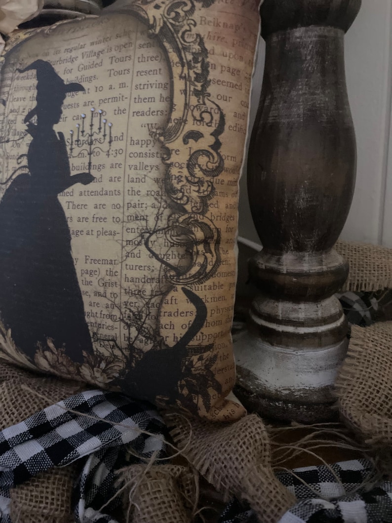 May include: A decorative pillow with a vintage aesthetic, featuring a black silhouette of a witch holding a candle. The pillow has a background of aged paper with text and is surrounded by burlap and a black and white checkered ribbon.