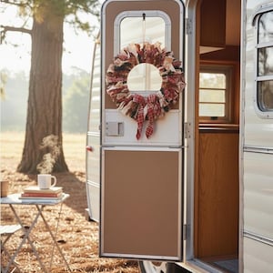 May include: A fabric rag wreath featuring a mix of red, white, and dark green plaid and solid strips of cloth tied together in a circular shape, hanging on the door of a white vintage travel trailer with a red plaid ribbon bow at the bottom.
