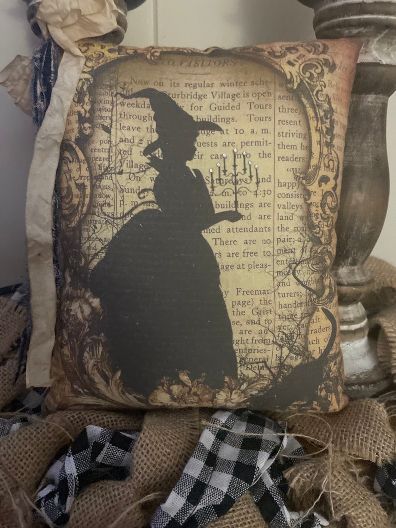 May include: Decorative pillow with a black silhouette of a witch on a vintage book page background. The pillow is square and has a distressed, antique look, perfect for Halloween decor.