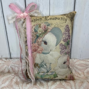 May include: A decorative pillow with a vintage Easter tea party design featuring two bunnies, flowers, and a teapot. The pillow is trimmed with pink lace and a pink ribbon.