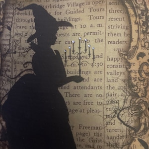 May include: Decorative pillow featuring a black silhouette of a witch holding a candelabra. The background is a vintage newspaper print with text and a decorative frame. Embellished with small, sparkling accents.