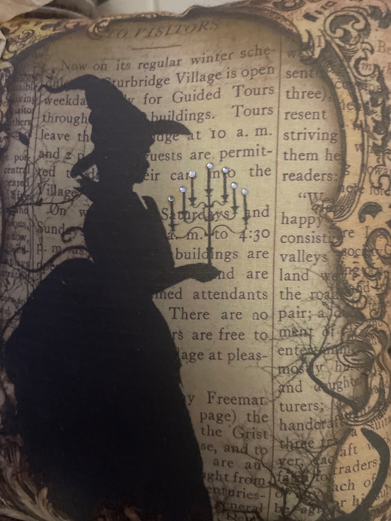 May include: Decorative pillow featuring a black silhouette of a witch holding a candelabra. The background is a vintage newspaper print with text and a decorative frame. Embellished with small, sparkling accents.